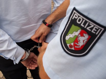 Polizist öffnet Handschellen an einer Person; Polizeilogo von NRW ist präsent im Bild.